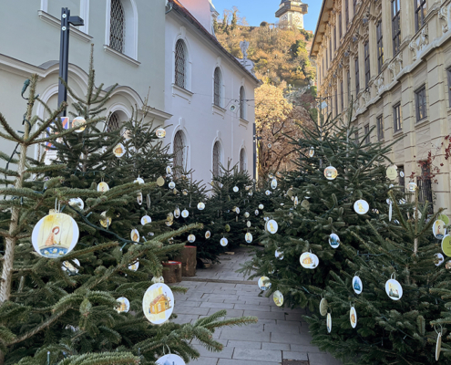 Tannenbäume mit illustrierten Anhängern stehen auf einem urbanen Platz, von Häusern eingerahmt, im Hintergrund ist der Schloßberg mit Uhrturm zu sehen