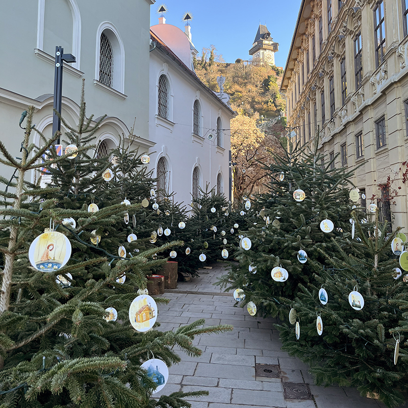 Tannenbäume mit illustrierten Anhängern stehen auf einem urbanen Platz, von Häusern eingerahmt, im Hintergrund ist der Schloßberg mit Uhrturm zu sehen