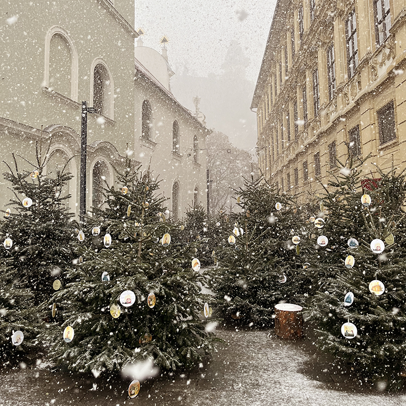 Schneetreiben, Tannenbäume mit illustrierten Anhängern stehen auf einem urbanen Platz, von Häusern eingerahmt, im Hintergrund ist der Schloßberg mit Uhrturm zu sehen