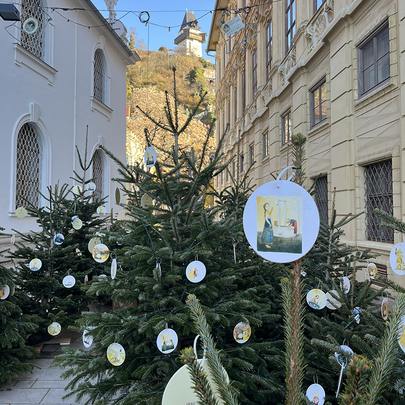 Tannenbäume mit illustrierten Anhängern stehen auf einem urbanen Platz, von Häusern eingerahmt, im Hintergrund ist der Schloßberg mit Uhrturm zu sehen