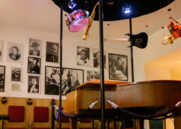 Detailaufnahme im Salon Stolz im Bereich der Lebensbühne. In der Mitte des Raumes steht ein Klavier von der Decke hängen Instrumente. An der Wand im Hintergrund hängen Fotografien von Robert Stolz
